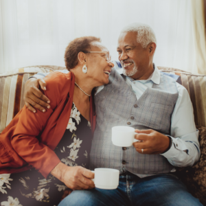An elderly couple holding coffee mugs smiling and hugging on a couch. 