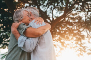 Picture of an older woman with glasses hugging an older man while standing under a tree.